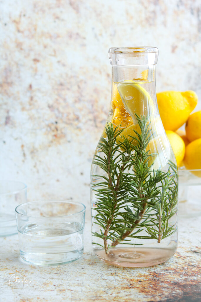 rosemary lemon water in carafe with lemons in the back with glasses 