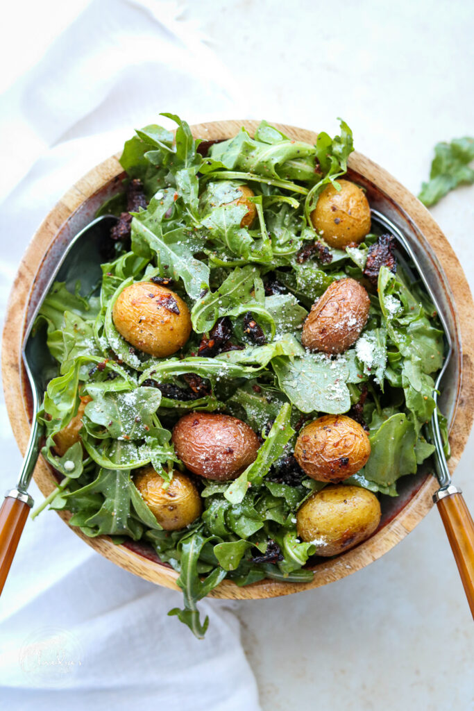 cooked potatoes with sun dried tomatoes and arugula in bowl with serving utensils
