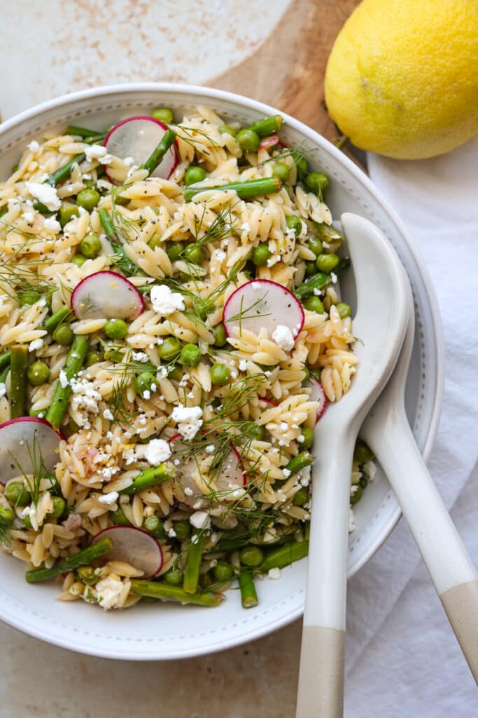 orzo salad with asparagus and peas in white bowl with serving utensils and lemon 
