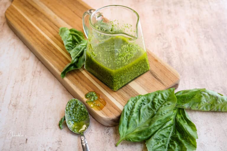 horizontal image of basil pesto vinaigrette with fresh basil and a spoon with the dressing on it on a small board.